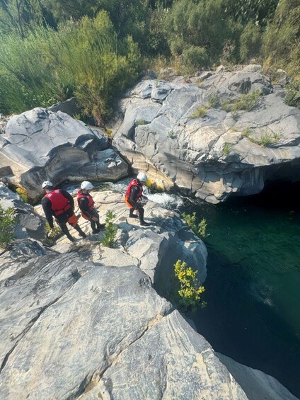 Three people in red jackets and helmets on rocky terrain near a body of water. 2