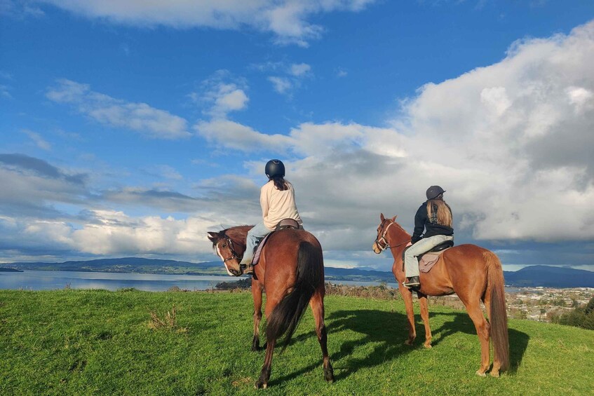 Two people riding horses on a grassy hill with a lake and mountains in the background. 2
