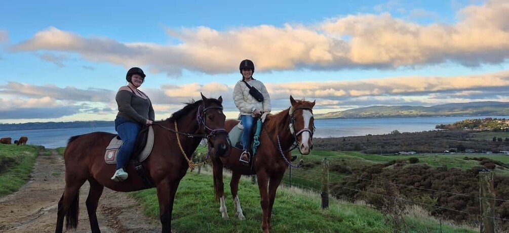 Two people riding horses on a dirt path with a scenic view of a lake and hills in the background. 3