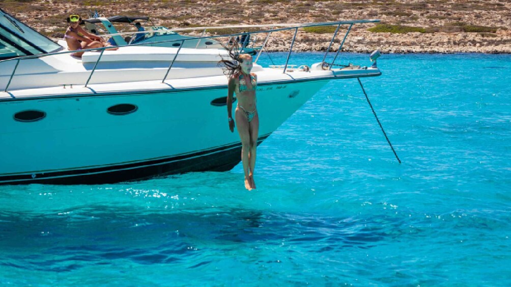 A boat with a person standing on the edge, another person sitting on the deck, and a rocky shoreline in the background. 1