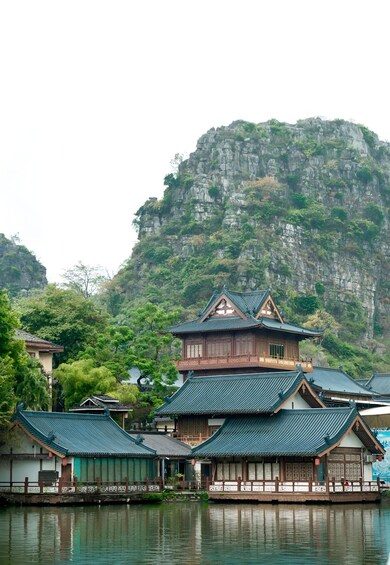 Traditional wooden buildings with green roofs by a calm body of water, surrounded by lush greenery and a rocky cliff. 4