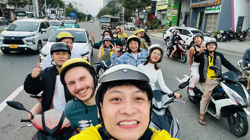 A group of people on scooters on a busy street with buildings and parked cars. 1