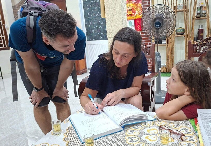 A person writing in a notebook at a table with a patterned tablecloth, a fan, and a decorative shelf in the background. 2