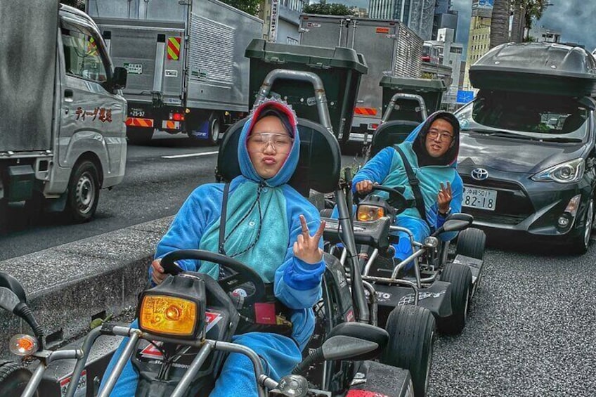 Two individuals in blue outfits driving go-karts on a city street with vehicles and buildings in the background. 1