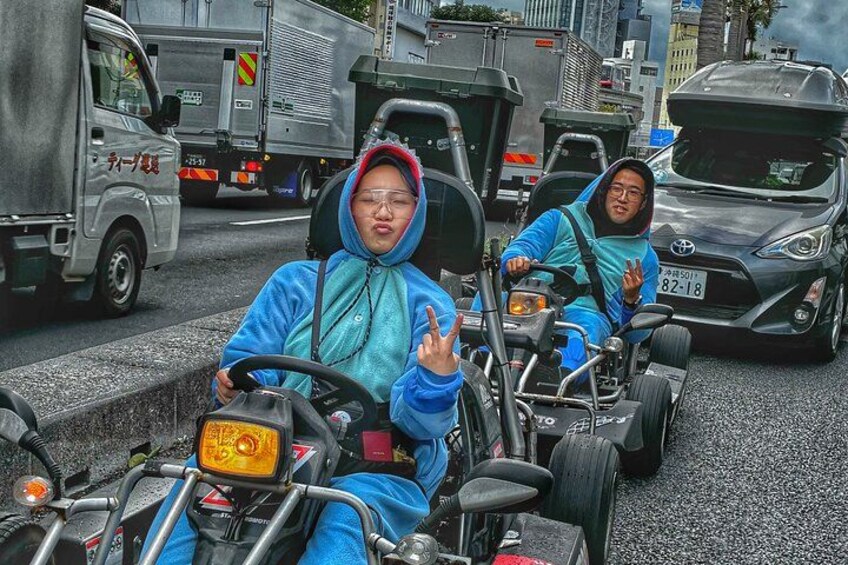 Two people in blue outfits driving go-karts on a city street with vehicles and buildings in the background. 1
