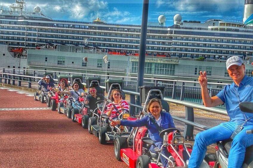 A group of people on go-karts lined up on a track with a cruise ship in the background. 4