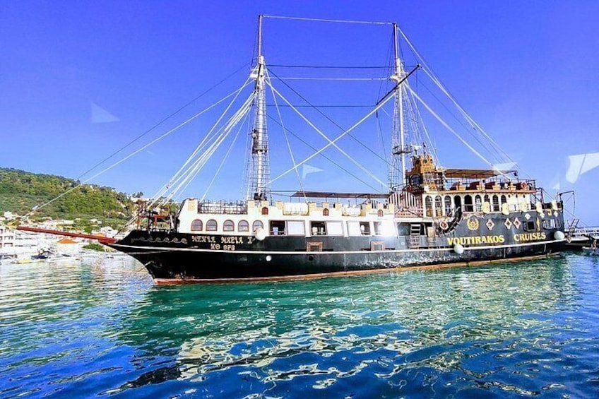 A large sailing ship docked at a harbor with clear blue water and a mountainous landscape in the background. 1