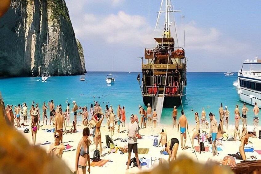 A crowded beach with people sunbathing and a large boat anchored near a cliff. 2