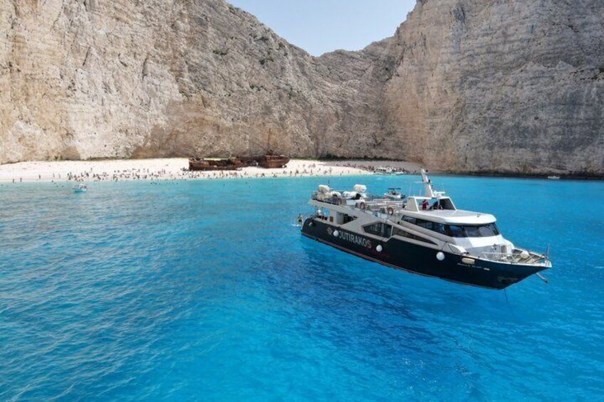 A boat named 'OUTRAKOS' on clear blue waters with a rocky coastline in the background. 1