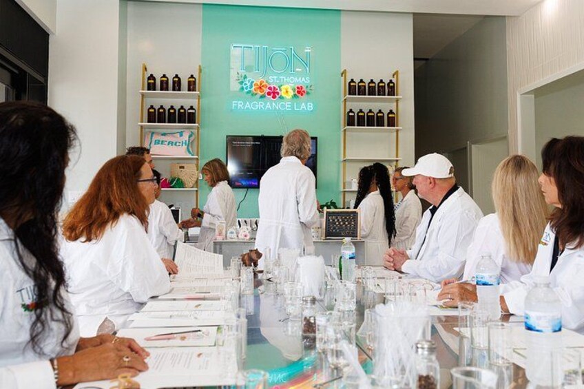 A group of people in white lab coats at a table with scientific equipment, in a room with shelves of bottles and a sign reading 'Tijon St. Thomas Fragrance Lab'. 5