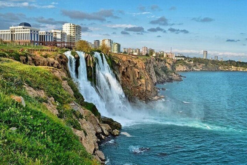 A coastal cityscape with a prominent waterfall cascading into the sea, surrounded by greenery and urban buildings. 1