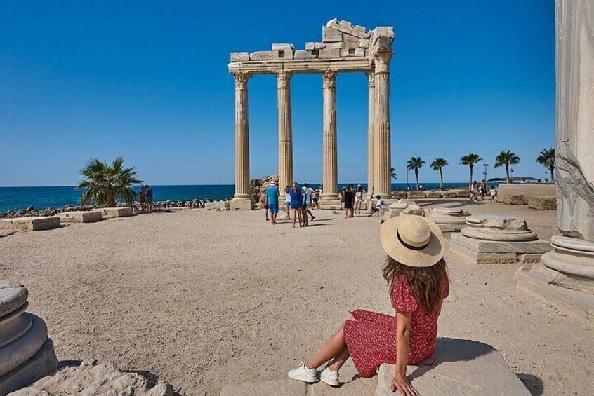 A woman in a red dress and hat sitting on a stone ledge, overlooking ancient columns and the sea. 1