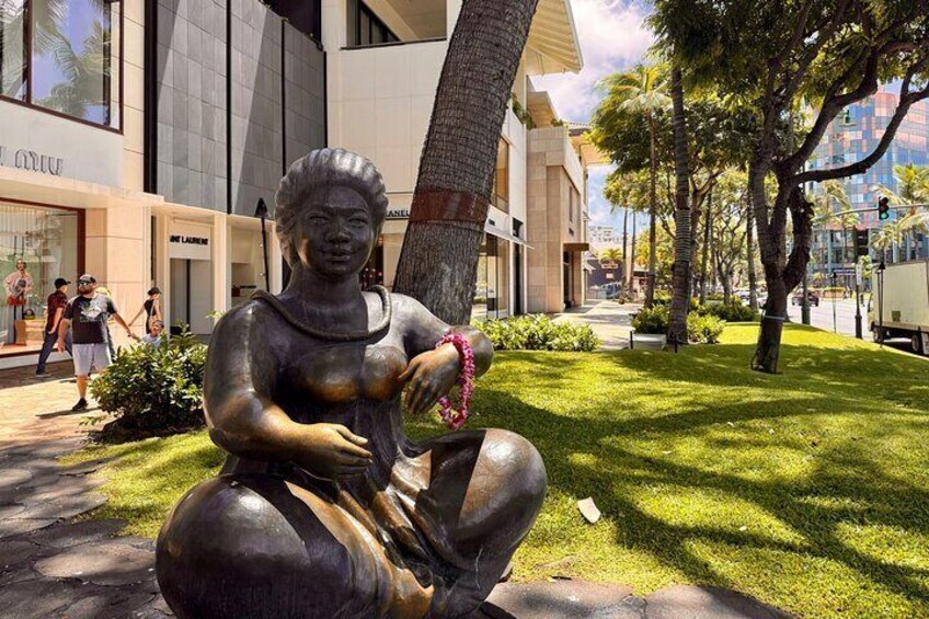 A bronze statue of a seated woman with a pink bracelet, located in a grassy area with buildings and pedestrians in the background. 4