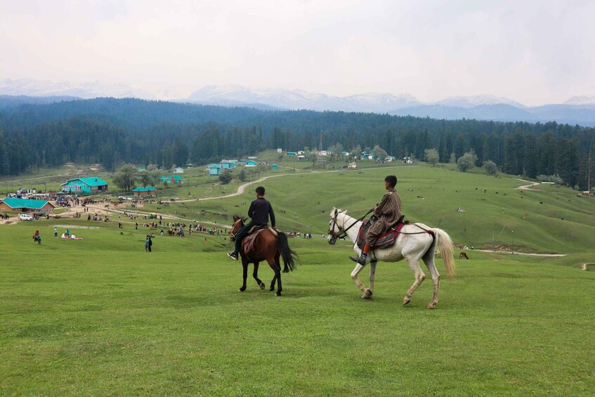 Two people riding horses on a grassy hill with a village and mountains in the background. 1