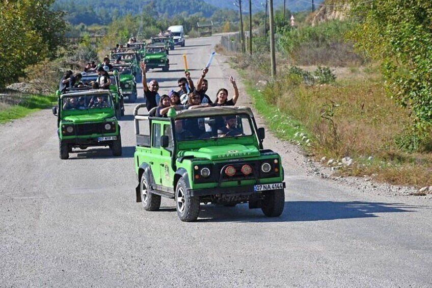 A convoy of green off-road vehicles on a rural road with passengers waving. 5