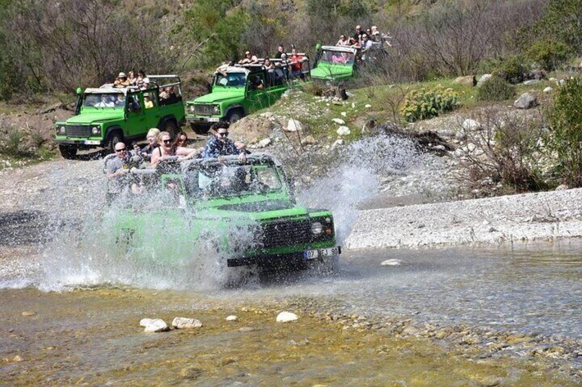 A green off-road vehicle driving through a shallow water crossing, with spectators watching from the side. 1
