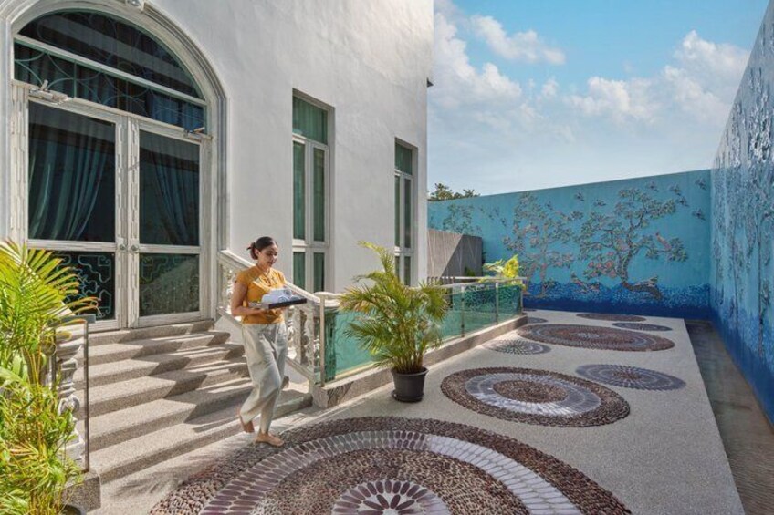 A person carrying a tray outside a building with a mural, a staircase, and potted plants. 2