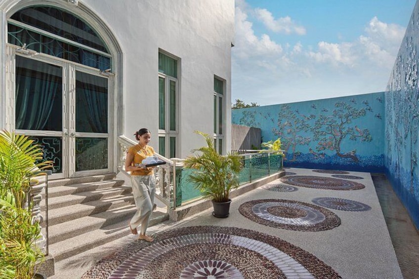 A person carrying a tray outside a building with a mural, a staircase, and potted plants. 2