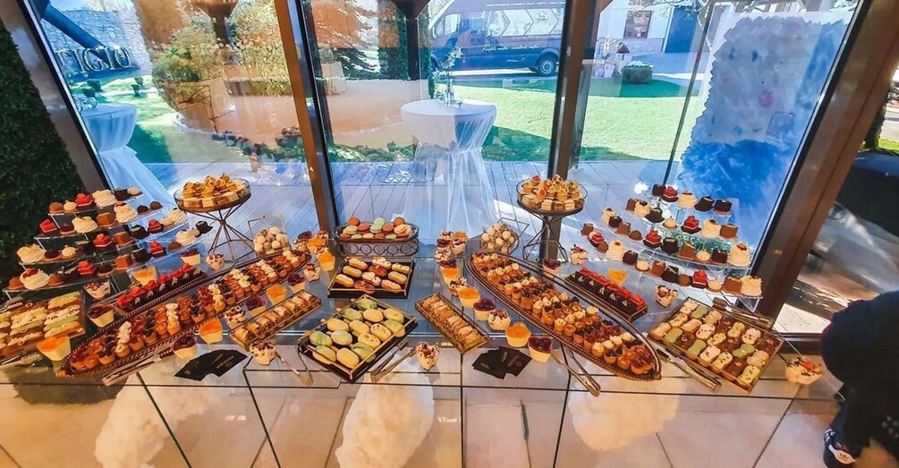 A variety of pastries and desserts displayed on multiple stands inside a building with large windows. 3