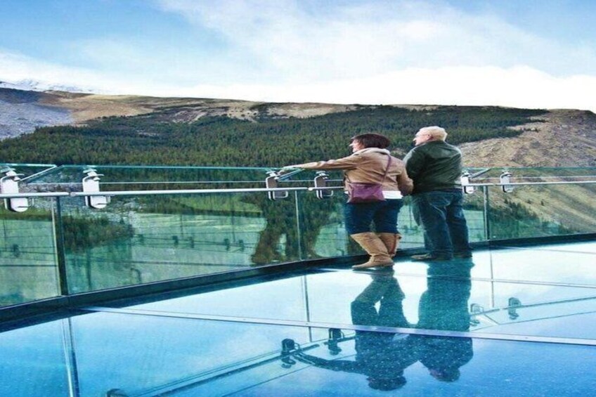 A couple standing on a glass floor balcony overlooking a mountainous landscape. 4