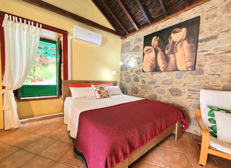 A bedroom with a stone wall, a bed with a red blanket, a wooden chair, and a window with curtains. 5