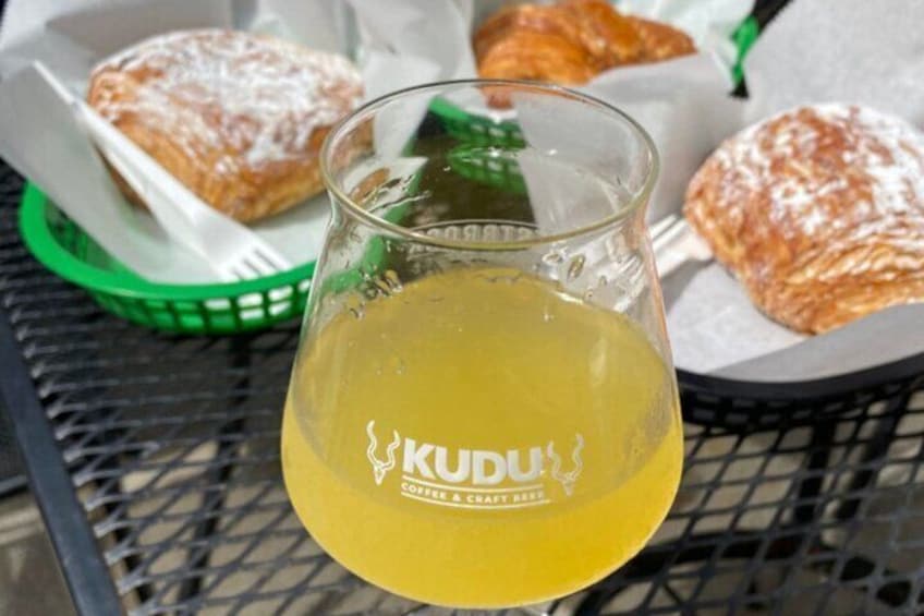 A glass of yellow liquid with 'KUDU Coffee Craft Beer' branding, pastries, and a green basket on a metal table. 2