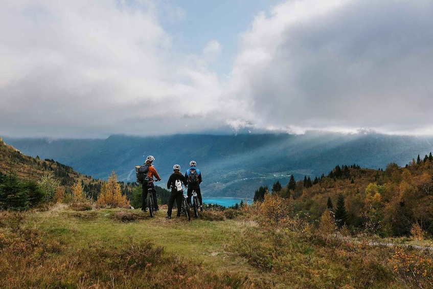 Three cyclists on a mountain trail with a lake and forested hills in the background. 2