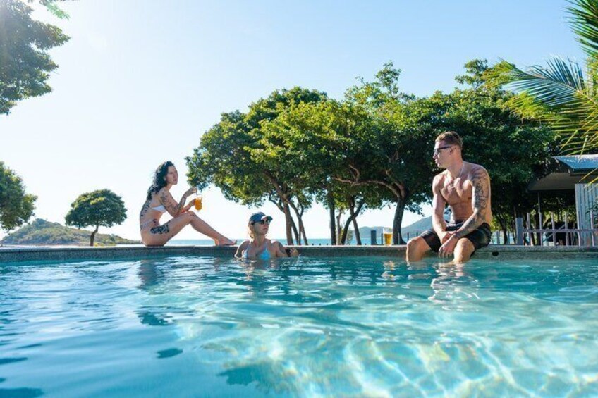 A swimming pool with people relaxing, surrounded by trees and a clear sky. 4