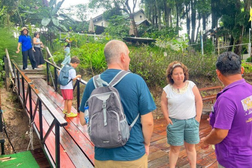 People on a wooden bridge in a tropical setting with greenery and a house in the background. 3