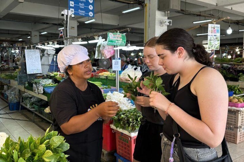 A person in a white chef's hat and black apron, holding a plant, standing next to a woman in a black tank top, both examining a plant in a grocery store. 4