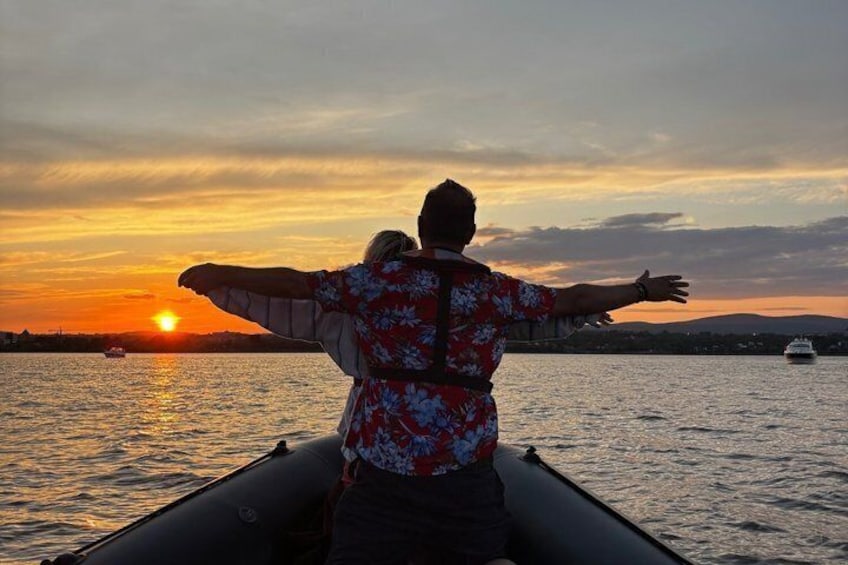 A person on a boat with arms outstretched, facing a sunset over water. 2