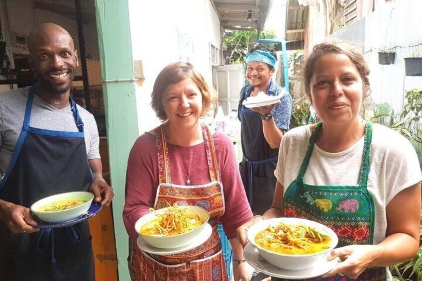 Three people holding bowls of food, wearing aprons, in an outdoor setting with buildings in the background. 1