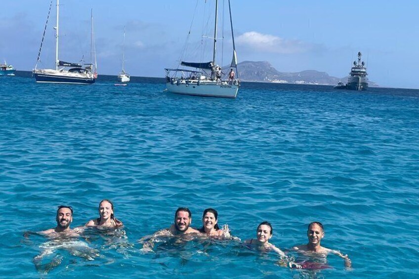 A group of people swimming in clear blue water with sailboats and a lighthouse in the background. 3