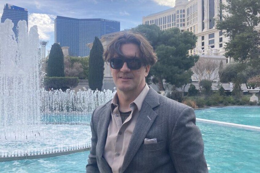 A person standing in front of a fountain with a pool, buildings, and trees in the background. 3