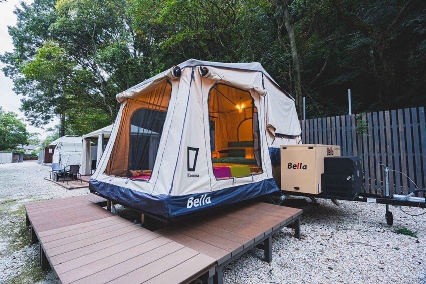A Bella tent set up on a gravel area with a wooden deck, surrounded by trees and a wooden fence. 3