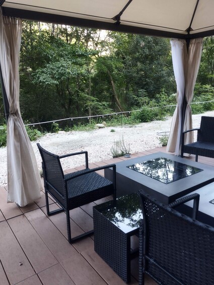 An outdoor patio with wicker furniture and a glass-top table, surrounded by a covered area with curtains, set against a backdrop of trees and gravel. 4