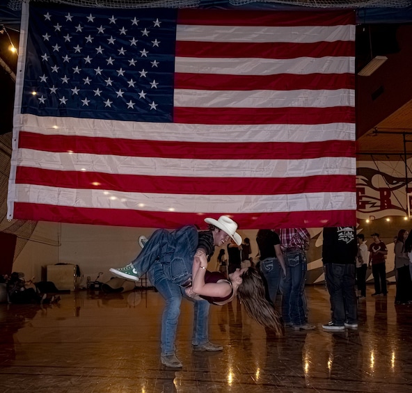 A large American flag, a person in a cowboy hat, and a crowd in a room with a wooden floor. 2
