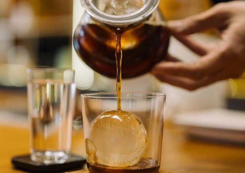 A person pouring a dark liquid into a glass with an ice sphere. 1