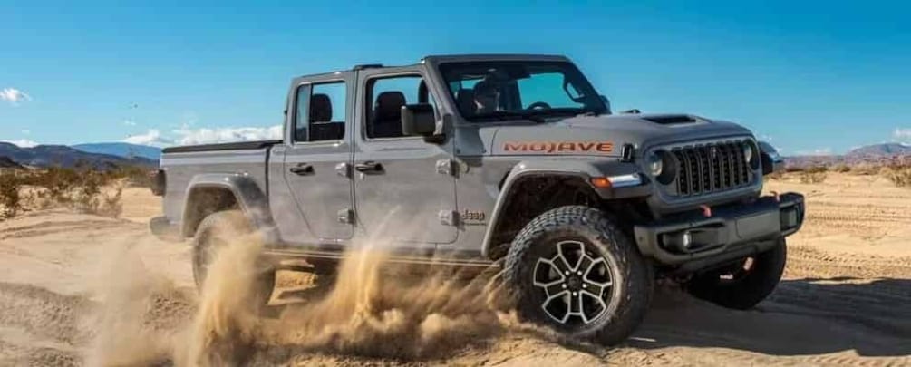 A gray off-road vehicle with 'MOJAVE' written on the side, driving through a desert with sand being kicked up. 1