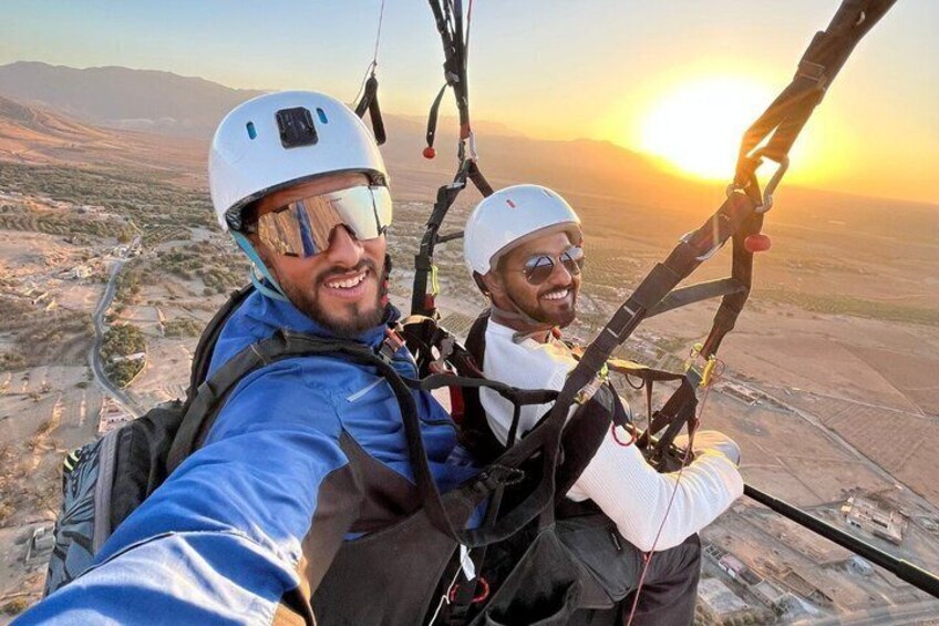 Two people in a harness with a parachute, taking a selfie with a sunset in the background. 1