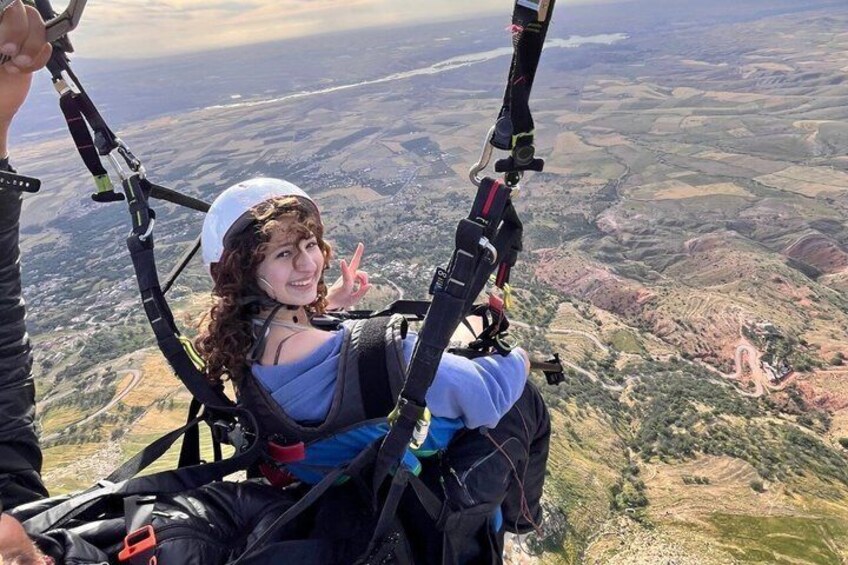 A person in a harness attached to a paraglider, with a landscape of rolling hills and a winding road below. 2