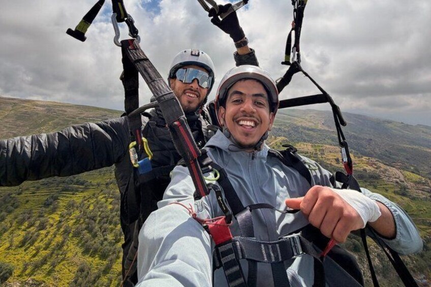 Two people in a tandem paragliding harness, with a mountainous landscape in the background. 4