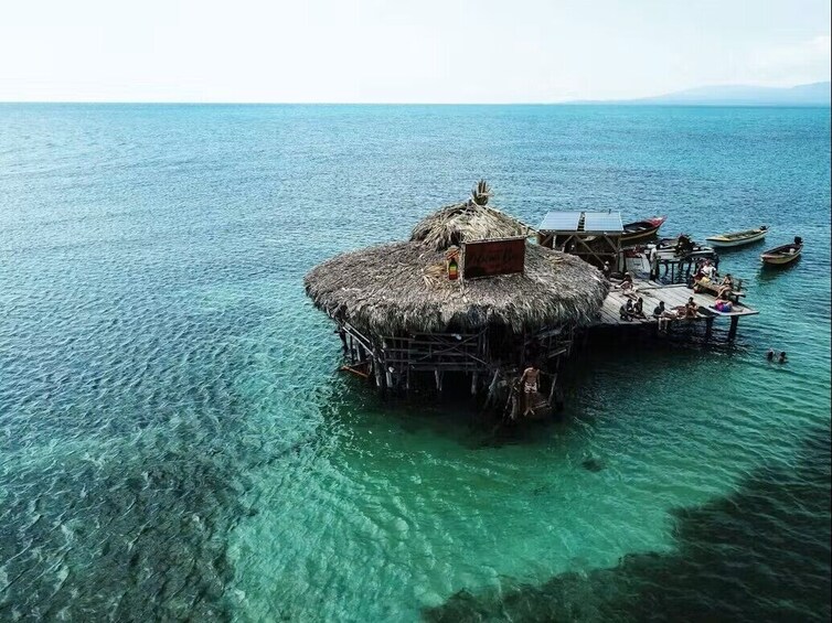 Une hutte à toit de chaume, construite sur pilotis, se dresse au milieu d’une eau turquoise et cristalline. 1