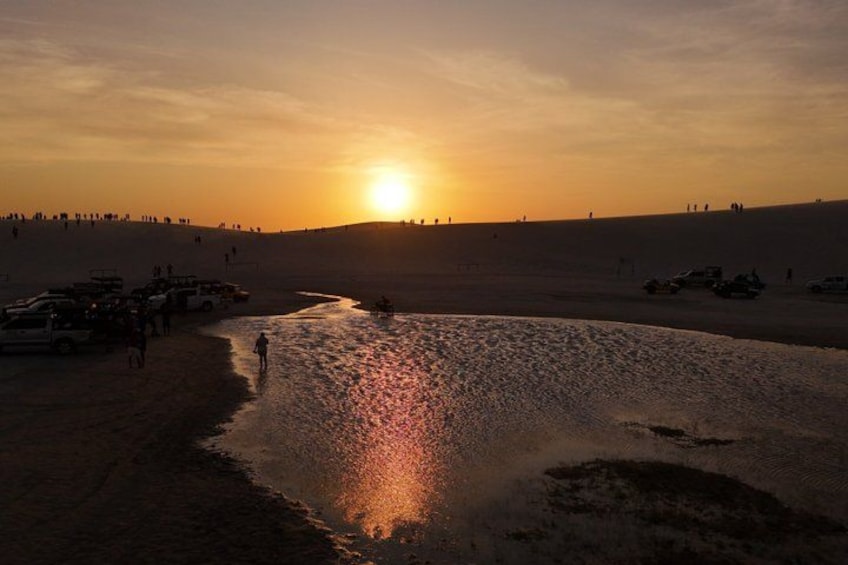A sunset over a desert landscape with a reflective water body, vehicles, and people. 2