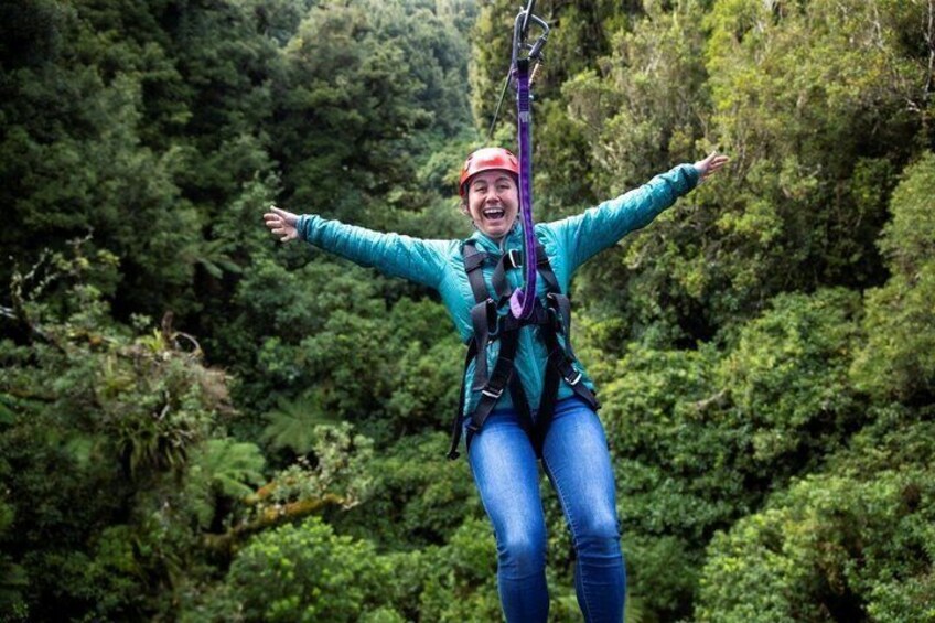 A person suspended in mid-air, wearing a harness and safety gear, with outstretched arms, against a backdrop of dense green foliage. 1