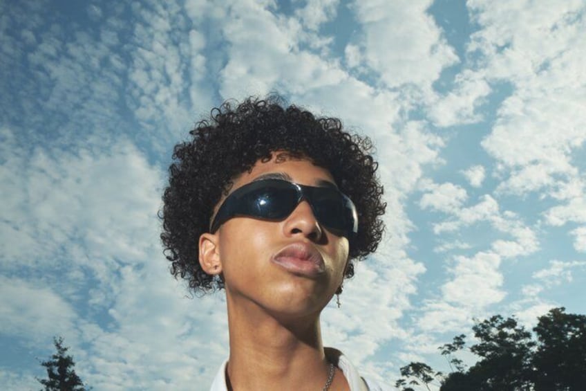 A person with curly hair and sunglasses against a sky with clouds. 4