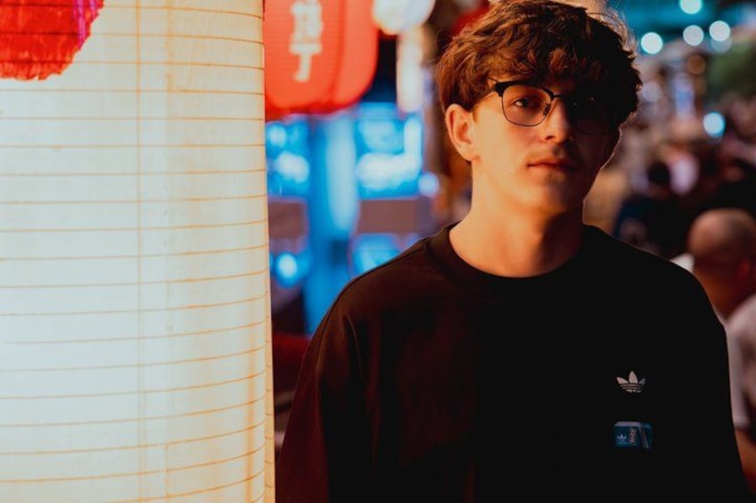 A person standing in a busy urban environment with neon signs and a red lantern in the background. 2