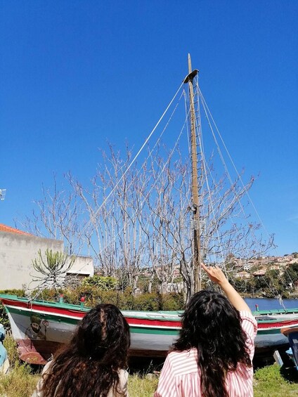 Two people observing a tall, slender pole structure with multiple ropes against a clear blue sky, with boats and trees in the foreground. 2