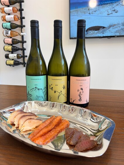 A plate with sliced meat and a glass dish on a table, with three bottles of wine in the background. 3