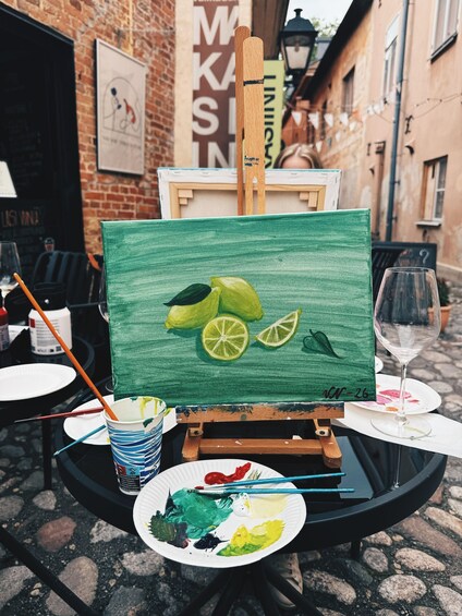 An easel with a painting of limes on a table, surrounded by art supplies and a wine glass. 3